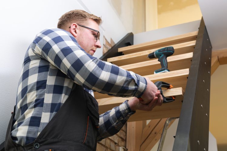 Man wearing a blue plaid shirt and safety glasses using a power drill on a wooden staircase during a home improvement project